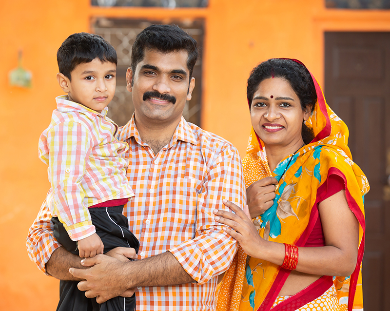 Portrait of young rural indian family standing outdoor at village house. Parent holding their little child, Mustache man wearing kurta and woman wear sari, Couple wearing traditional cloths with kid